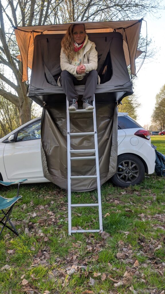 Blonde Frau sitzt auf Leiter eines Dachzelts auf weißem Auto