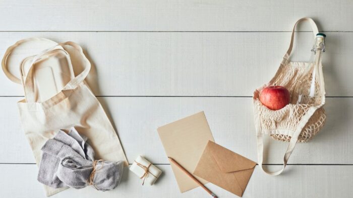 eine weiße Wand, an der ein Stoffbeutel hängt, daneben Seife, ein Briefumschlag und Stift, sowie ein gehäkelter Einkaufsbeutel mit einem Apfel und einer Flasche