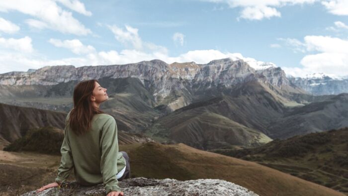 Frau sitzt auf Felsen vor bergigem Hintergrund. symbolisch für Ruhe und bewusste Atmung